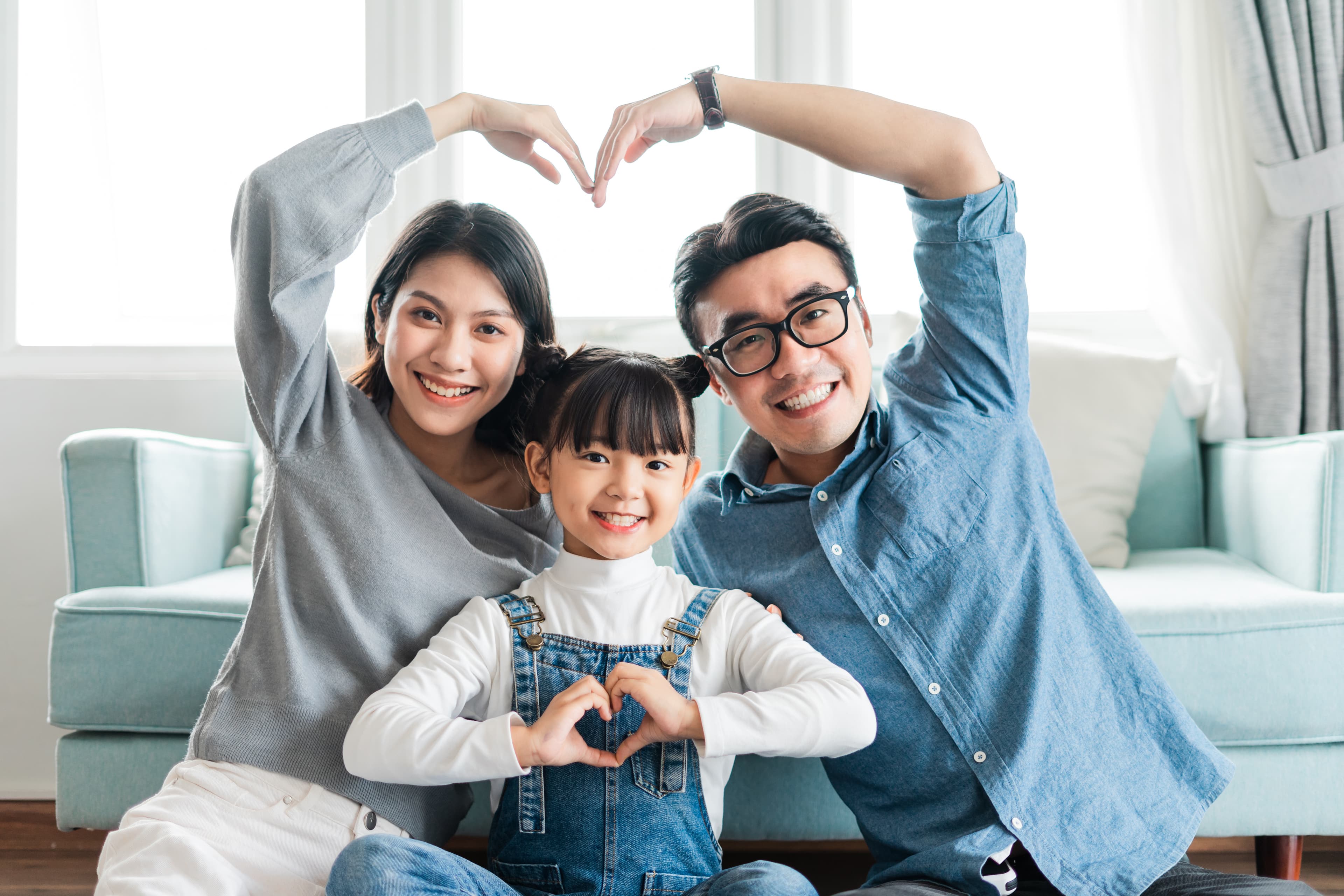 Cha mẹ và con gái ngồi cạnh nhau trong phòng khách, cùng chụm tay tạo hình trái tim